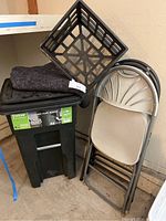 Photo of the 32 gallon Totter trashcan with a gray moving blanket, black plastic crate, and three beige folding chairs stacked against a wall in a garage.