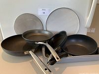 Four different sized non-stick fry pans, one a mini wok, arranged with two round metal splatter covers visible behind them.