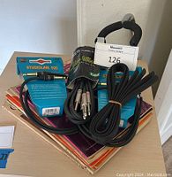 Stack of music books topped with guitar cords and a guitar strap, showing packaging and titles of books.