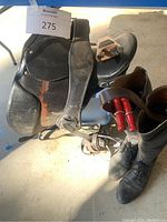 Photo showing black English leather saddle with attached stirrup leathers and metal stirrups alongside pair of men's black riding boots with red boot pullers.