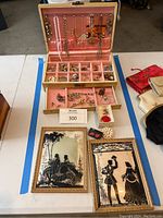 Jewelry box open showing necklaces hanging inside lid and assorted costume jewelry in compartments and drawers, with two framed silhouette art pieces and small accessories surrounding the box.