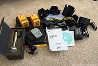 Photo showing two microphones (silver and black), Kodacolor-X film boxes, Canon battery charger, Nikon lens covers, camera strap, lens hoods, and photo paper pack.