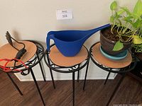 Three black metal plant stands with cork tops arranged side by side; the right has a live plant in a brown pot, the middle has a blue watering pitcher, the left has a slingshot with black and red accents