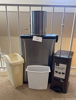 Front view of five trash cans: 2 metal step cans with lids, one round metal bin on top, and 2 plastic bins including yellow and white ones.