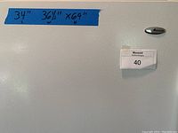Close up view of refrigerator door showing Amana logo, Maxsold tag, and appliance dimensions on blue tape