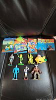 Overview of all figures and trading cards laid on a black surface. Eight small figures in bright colors with detailed character features, including blue, green, yellow, and brown characters. Several trading cards displayed above figures.