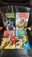 Four comic book issues displayed on a leather chair showing their front covers with vibrant colors and characters Cyclops and Phoenix.