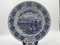 Front view of blue and white collector plate featuring pioneer scene and maple leaf border.