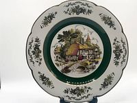 Front view of the decorative ceramic plate showing the village scene at center with green band and floral border with gold accents.