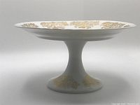 Front side view of the white porcelain cake stand showing hand-painted gold floral accents on rim and pedestal base.
