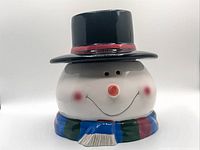 Front view of the ceramic snowman cookie jar showing the snowman's face, black top hat with red band, and colorful scarf base.