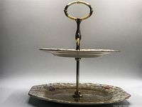 Side view of the vintage 2-tier tidbit stand showing lower and upper ceramic plates with floral pattern and gold trim, gold metal support and handle