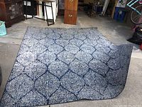 Full view of 8x10 feet blue patterned area rug. Rug lies partly folded in a grey and black speckled geometric pattern with darker blue outlines.