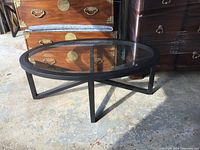 Oval glass top coffee table with black metal frame shown on a concrete floor next to wooden furniture