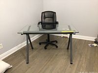 Full view of modern desk with clear glass top and black pleather chair behind it in a corner room with grey walls and wood floor.