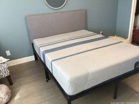 Full view of queen bed with grey upholstered headboard, black metal frame, and Sealy mattress with horizontal blue stripes on grey fabric.