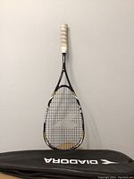 Squash racquet front view leaning against a wall with the Diadora black carrying case beneath it