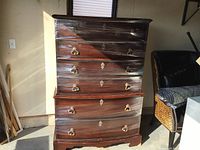 Front view of the tall boy dresser wrapped in plastic, showing multiple drawers with brass ring pulls and keyholes, dark wood finish.