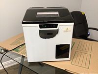 Front view of the white and black Vivo Home icemaker sitting on cardboard, showing peeling protective plastic on stainless steel panel and control buttons.