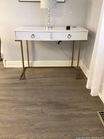 Full front view of white desk showing two drawers with gold ring handles and gold geometric legs. Small acrylic lamp and wireless charging pad on top.