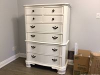 Front view of white highboy dresser with 7 drawers, ornate handles, curved skirt and decorative side carvings.