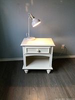 Creamy white nightstand with drawer and open shelf, lamp placed on top, showing overall setup