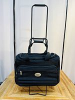 Front view of black American Tourister travel bag with extended trolley handle and front zippered pocket.