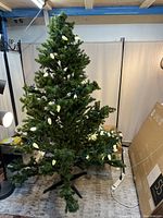 Artificial Christmas tree standing upright with Christmas lights wrapped around it, showing complete assembly and overall good condition. Box with packaging visible on the side.