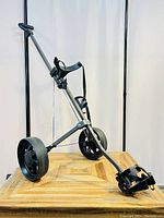 View of the folding golf pull cart showing metal frame, two large wheels, securing straps, and foldable design.