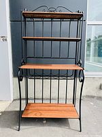 Front view of midcentury style baker's rack with black iron frame and wood shelves placed outdoors against a dark wall.