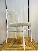 Front angle of white metal frame dining chair with beige textured cushioned seat and back on a wooden platform.