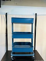 Full frontal view of the AGTEK 3-tier rolling utility cart, showing three metal storage trays in aristocratic blue with tubular frame and caster wheels.