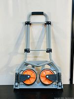 Front view of the VEVOR folding hand truck in folded position showing orange and black wheels and silver frame.