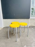 Photo showing three yellow round stools with curved silver metal legs standing on concrete against a dark wall.