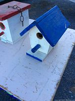 Photo of white bird house with blue roof, perched on a table with another bird house partially visible