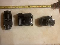 Photo shows Nikon MG-ES6000 film camera, Canon digital camera, and MZ Ricon 70-210mm telephoto zoom lens alongside a measuring tape for scale.