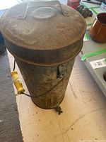 Photo showing a tall cylindrical metal can with a spout at the bottom and a yellow handle attached to a wire handle, surface rusted and weathered, lid present.