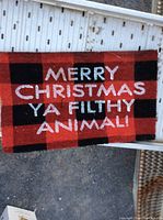Top view of rectangular welcome mat showing red and black checkered pattern with white text 'MERRY CHRISTMAS YA FILTHY ANIMAL!'