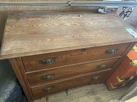 Front view of medium brown wooden dresser with three drawers showing ornate metal handles and visible wood grain.