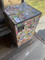 Photo showing top and part of one side of small fridge fully covered in colorful stickers placed on wooden platform.