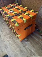 Full side view of orange trunk showing yellow metal hinges, corner brackets, and leather handle on side.