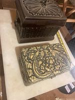 Two wooden keepsake boxes shown side by side on a light-colored surface, one dark carved wooden box and one lighter ornate floral box.