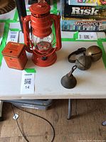 Photo showing red metal lantern, orange vintage tin container, and three bells (two large flat round bells on leather strap and one small bell) on white table with board games in background