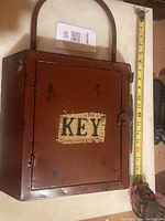 Closed rust-colored metal key holder box with 'KEY' patch and curved handle, measuring about 12 inches tall.