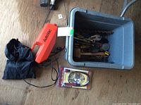 Photo shows a grey plastic bin filled with miscellaneous metal hand tools including wrenches, pliers, screwdrivers, a rubber mallet, and a large drill bit, next to an orange Autoloc handheld tool, a black pouch, and a packaged cable lock.