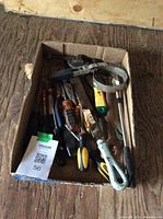 Photo showing a box with assorted screwdrivers, hand saw blade, pliers, and pipe clamp band.