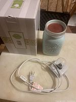 Blue Mason jar candle with reddish wax and metal lid, electric plug-in light bulb fixture with cord and switch, all placed on a table surface.