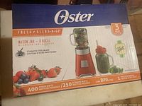 Front side of Oster Fresh Blend-N-Go boxed blender showing red base and glass mason jar blender container, emphasizing stainless steel blade and BPA free materials.