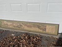 Wide shot of the framed long horizontal Asian scroll print leaning against a garage door outdoors on ground with dry leaves.