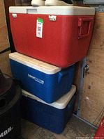 Three stacked coolers: a red and white Coleman cooler on top, a blue and white Coleman cooler in the middle, and a small solid blue cooler on the bottom